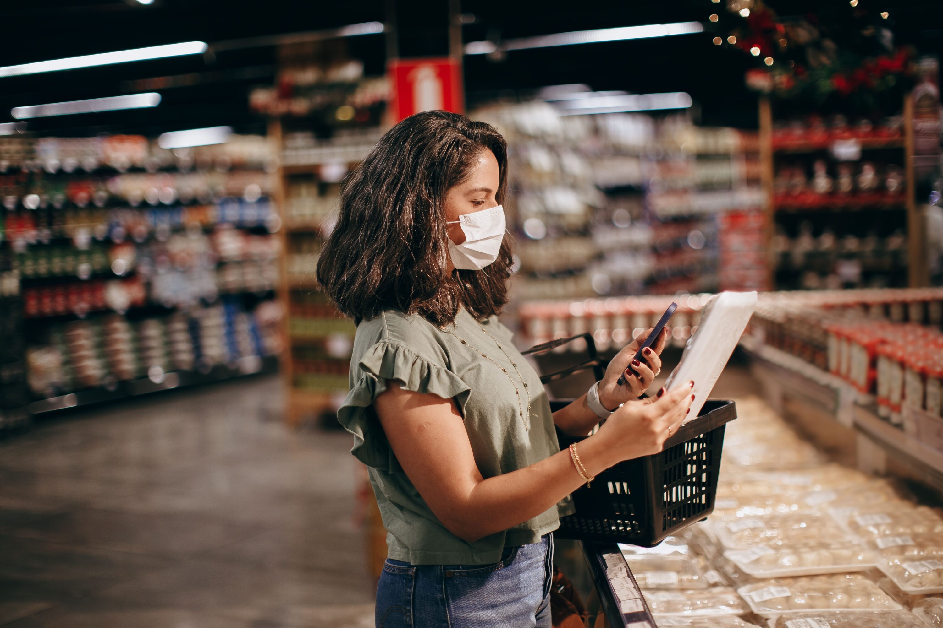 A woman checking her smartphone while shopping in a grocery store aisle, illustrating how retail media advertising connects digital intent with physical brick-and-mortar shopping.